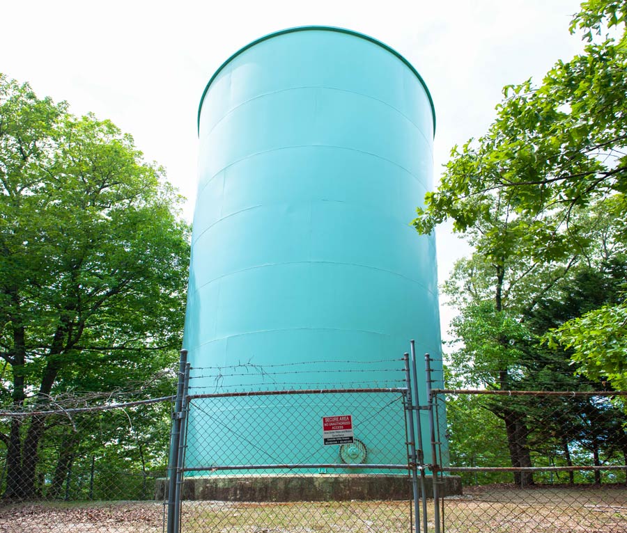 Large blue green water storage tank surrounded by trees and secured by a chain link fence
