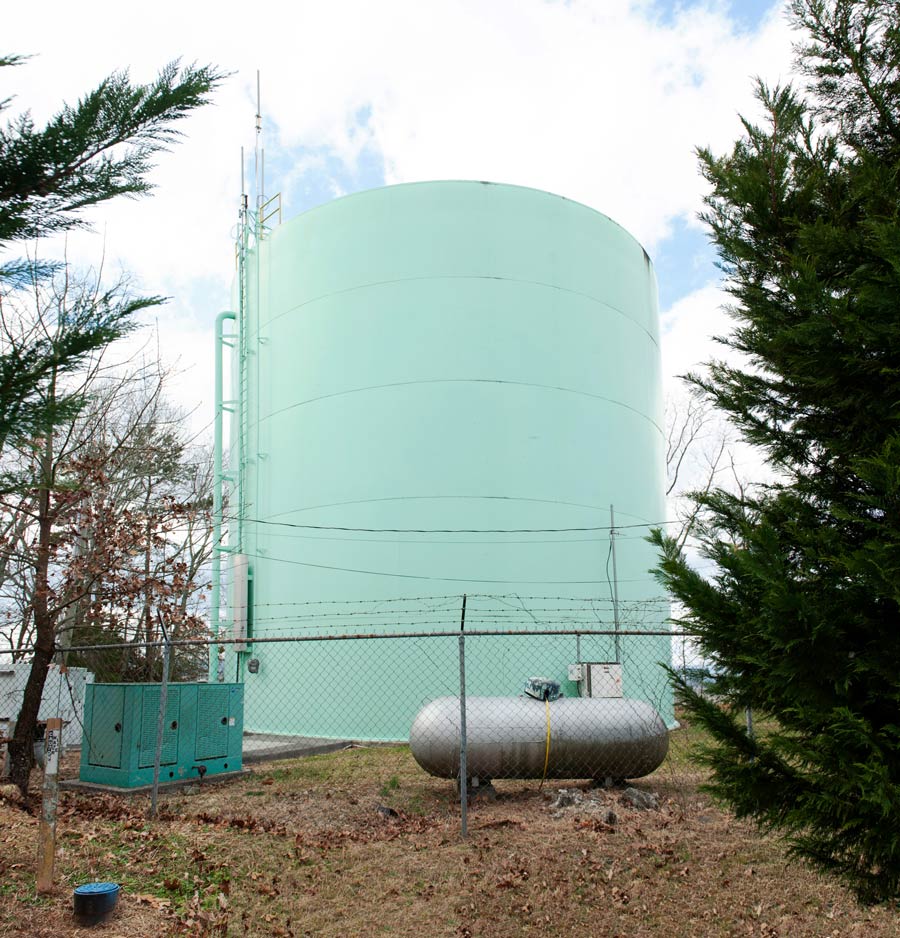 Large green elevated water storage tank with ladder access, fenced perimeter, and utility equipment at the base